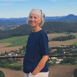 Frau mit weißen Haaren steht an einer Aussichtsplattform, Blick über Dorf, Felder und bewaldete Hügel unter blauem Himmel.