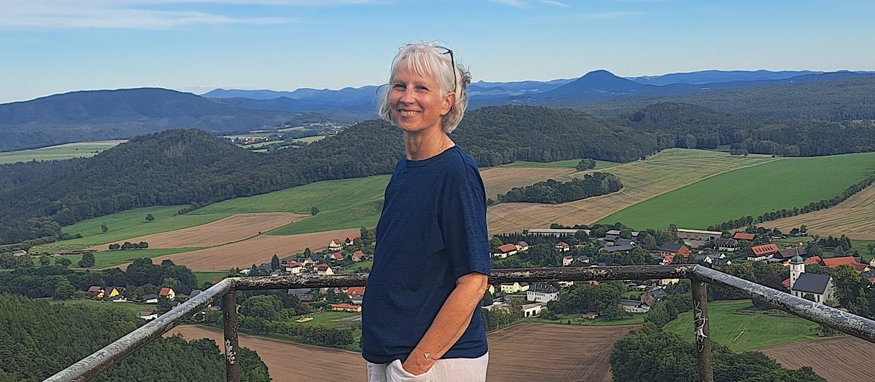 Frau mit weißen Haaren steht an einer Aussichtsplattform, Blick über Dorf, Felder und bewaldete Hügel unter blauem Himmel.