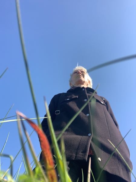 Focusing-Coach Birgit Krüger steht in schwarzem Mantel auf einer Wiese, von unten fotografiert – Grashalme im Vordergrund, weiter Himmel über ihr.