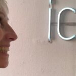 Frau mit weißem Haar blickt unsicher auf leuchtenden Schriftzug ‚Ich‘ an einer weißen Wand.