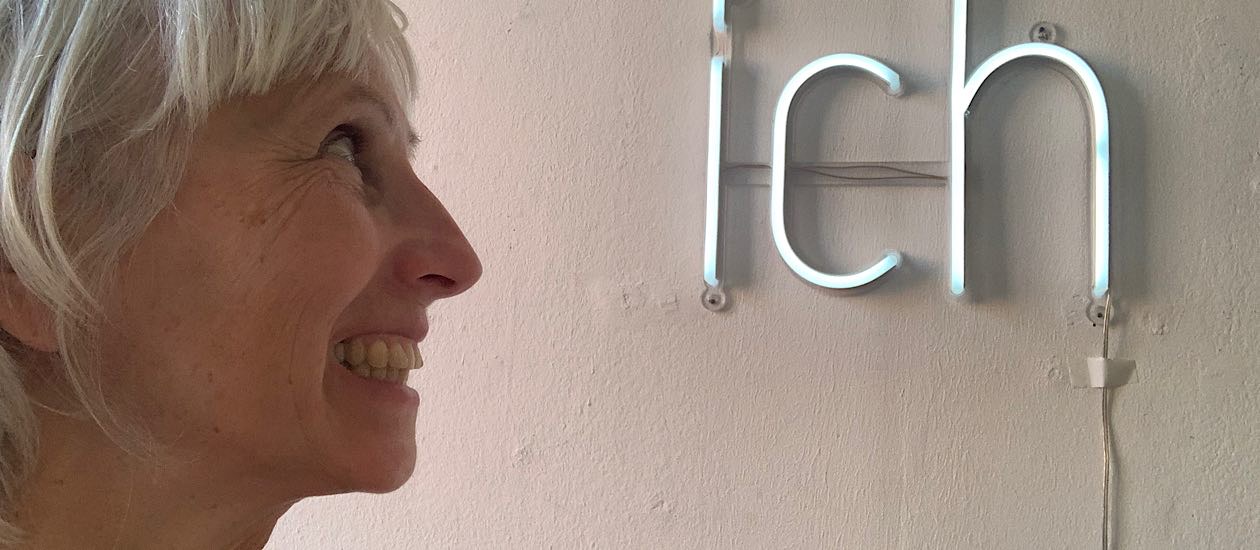 Frau mit weißem Haar blickt unsicher auf leuchtenden Schriftzug ‚Ich‘ an einer weißen Wand.