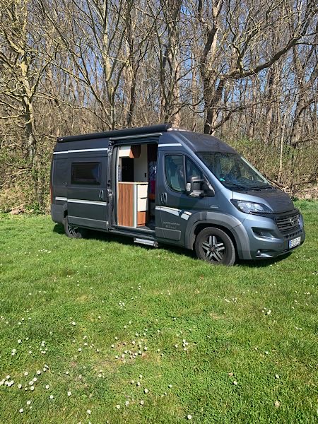 Grauer Campervan steht mit geöffneter Schiebetür auf einer grünen Wiese vor einem Wald. Er gehört Focusing-Coach Birgit Krüger