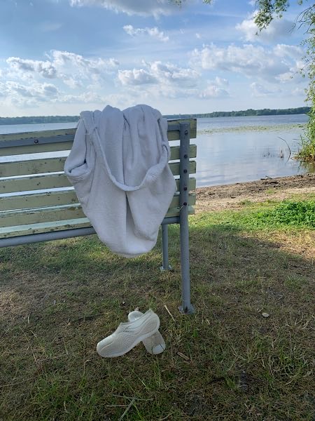 Weißer Bademantel auf Bank am Seeufer, darunter Badeschuhe im Gras; Blick auf ruhigen See und blauen Himmel mit Wolken.