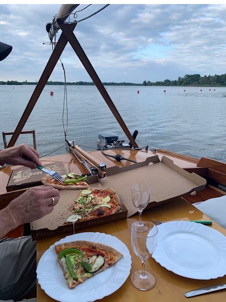 Pizzaessen auf einem Segelboot bei ruhigem Wasser; zwei Teller, Gläser und Pizzaschachtel auf Holztisch mit Blick auf den See.