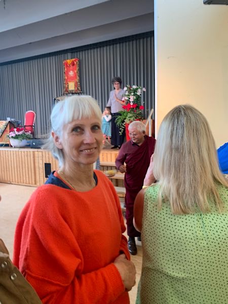 Birgit Krüger in orangem Pullover lächelt in die Kamera, im Hintergrund tibetischer Mönch bei Veranstaltung mit Blumen und Bühne.
