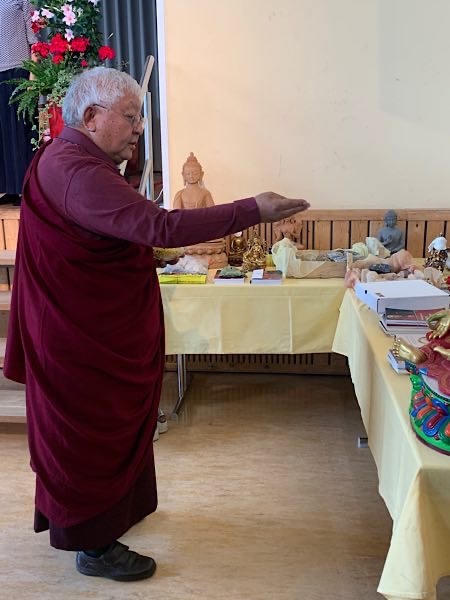 Älterer buddhistischer Lehrer in roter Robe segnet rituelle Gegenstände auf einem Tisch mit Figuren und Texten.