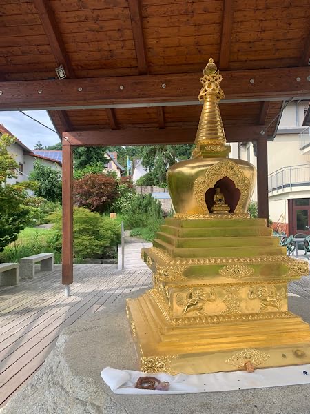 Goldener buddhistischer Stupa unter Holzpavillon in gepflegtem Garten, mit kleiner Buddhafigur in der Nische.