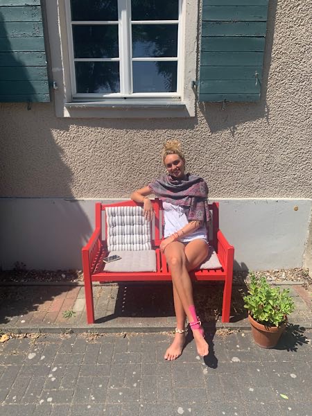 Frau sitzt entspannt auf roter Bank vor einem Haus mit Fensterläden, barfuß, sommerlich gekleidet, sonniger Tag, Pflanze daneben.