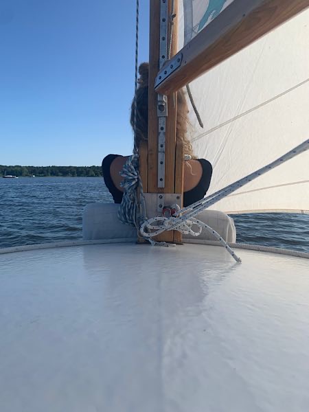 Frau sitzt vorn auf einem Segelboot, Rücken zur Kamera, Blick auf See und Horizont, Segel und Seile im Vordergrund sichtbar.
