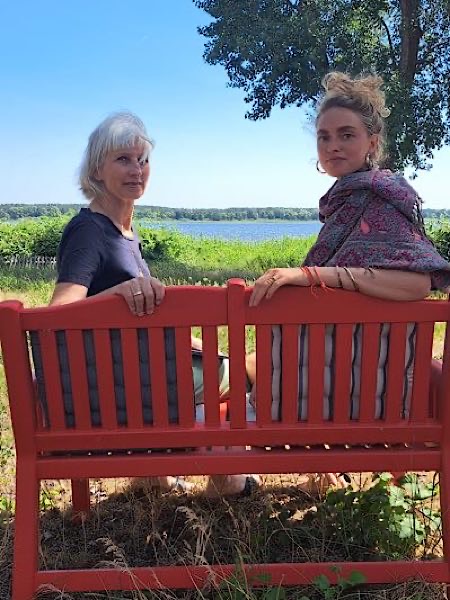 Birgit Krüger sitzt mit junger Frau auf roter Bank am See, blicken zur Kamera; sommerliche Landschaft mit Wasser, Wiese und Baum im Hintergrund.