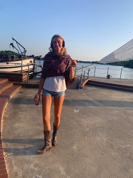 Frau mit Kopfhörern und Drink in der Hand im Abendlicht bei SIlent Disco am Wasser, leger gekleidet, Boot und Steg im Hintergrund.