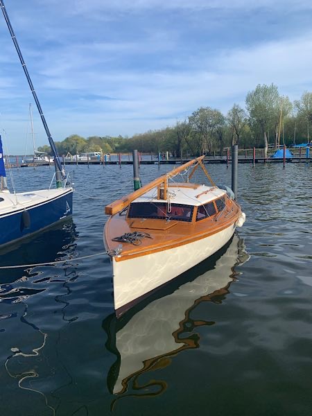 15er-Jollenkreuzer mit weißem Rumpf und Holzdeck liegt im Hafen, gespiegelt im Wasser; ruhige Stimmung, Bäume und Segelboote im Hintergrund.