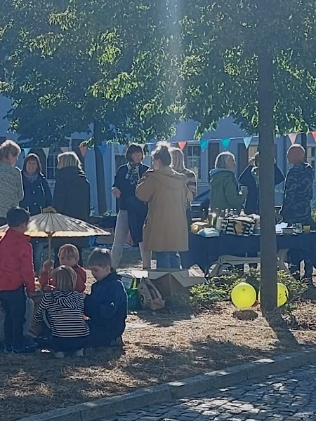 Menschen beim Tag der Nachbarschaft im Schatten von Bäumen; Kinder spielen im Vordergrund, Erwachsene stehen an einem bunten Stand.