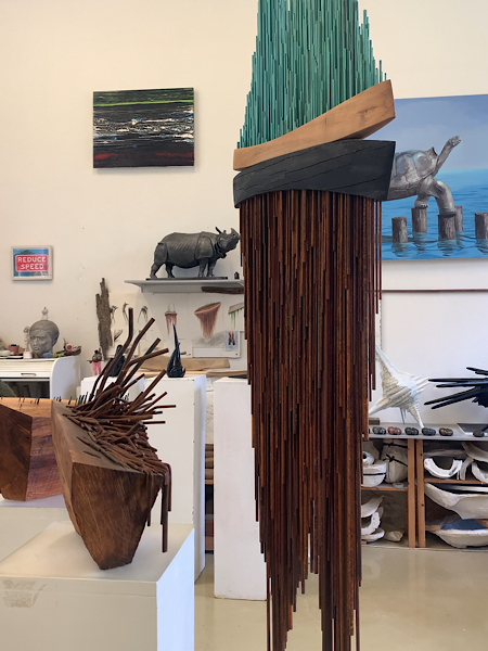Raum mit modernen Holzskulpturen; im Zentrum eine hohe Skulptur aus Stäben, grünem Element und Holz. Bilder und Figuren im Hintergrund.