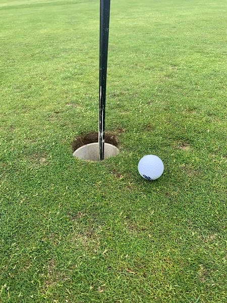 Golfball kurz vor dem Einlochen, direkt am Loch mit Fahnenstange; grüne, gepflegte Rasenfläche auf dem Golfplatz.