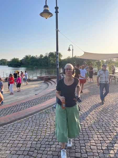Birgit Krüger mit Kopfhörern bei SIlent Disco, grüner Hose und Getränk an der Uferpromenade entlang, Menschen im Hintergrund.