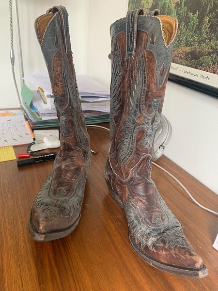 Cowboystiefel in braun und grau auf Schreibtisch, im Hintergrund Büromaterial