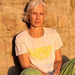 Focusing-Coach Birgit Krüger sitzt in weißem T-Shirt und grüner Hose im warmen Abendlicht vor einer Sandsteinwand und blickt ruhig und offen in die Kamera