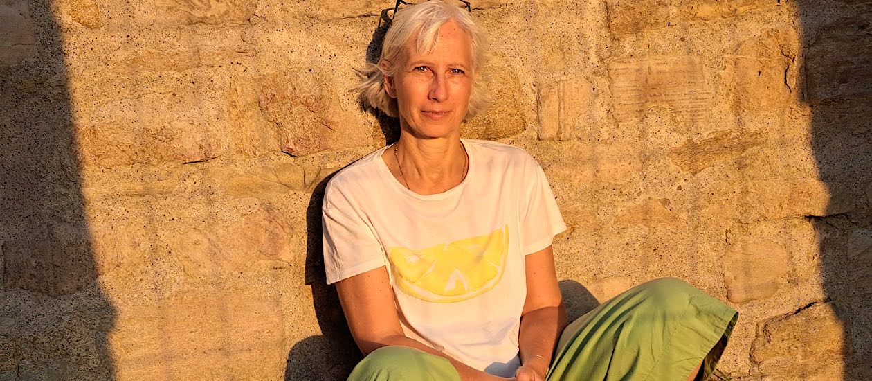 Focusing-Coach Birgit Krüger sitzt in weißem T-Shirt und grüner Hose im warmen Abendlicht vor einer Sandsteinwand und blickt ruhig und offen in die Kamera