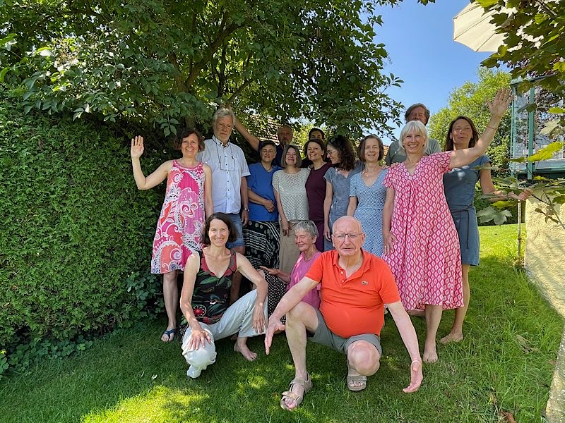 Gruppenfoto im Garten bei Sonnenschein: 15 fröhliche Menschen, teils stehend, teils hockend, winken und lachen in die Kamera.