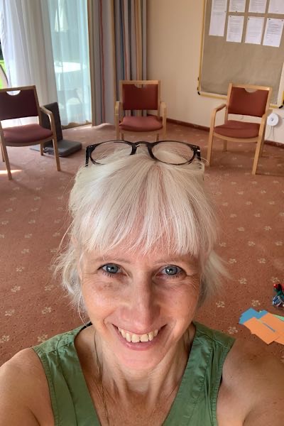 Focusing-Coach Birgit Krüger lächelt in die Kamera, im Hintergrund ein Seminarraum mit Stühlen und Moderationsmaterial auf dem Boden.