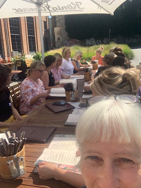 Birgit Krüger mit einer großen Frauengruppe beim Mittagessen draußen an einem langen Holztisch unter Sonnenschirmen in urbaner Umgebung.