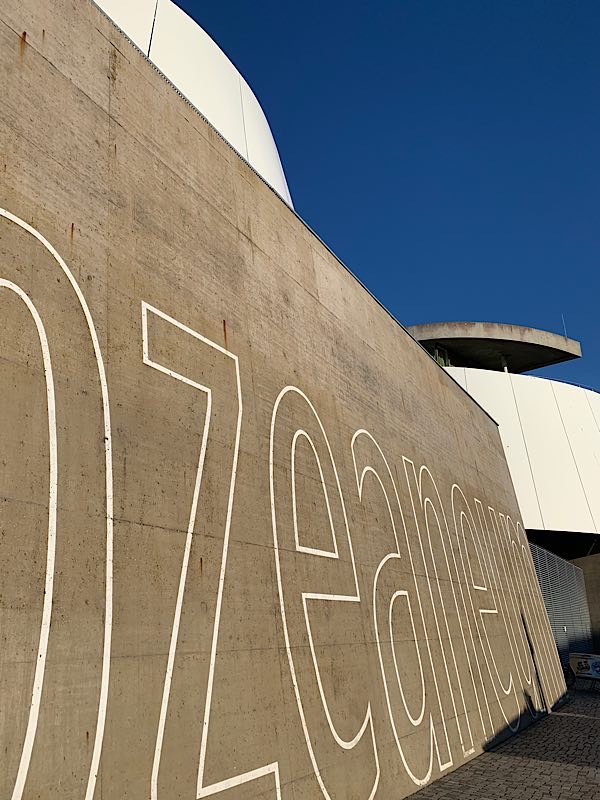 Betonwand mit großer weißer Schrift „Ozeaneum“ in Stralsund, fotografiert aus steilem Winkel bei blauem Himmel.