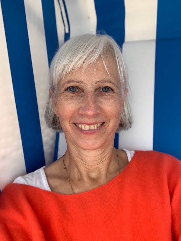 Birgit Krüger mit weißem Haar und rotem Pullover sitzt lächelnd in einem blau-weiß gestreiften Strandkorb und schaut in die Kamera.
