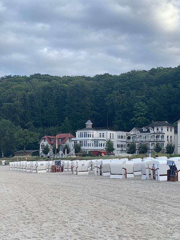 Weiße Strandkörbe am Sandstrand vor historischen Villen von Binz auf Rügen, dahinter dichter Wald unter wolkigem Himmel.