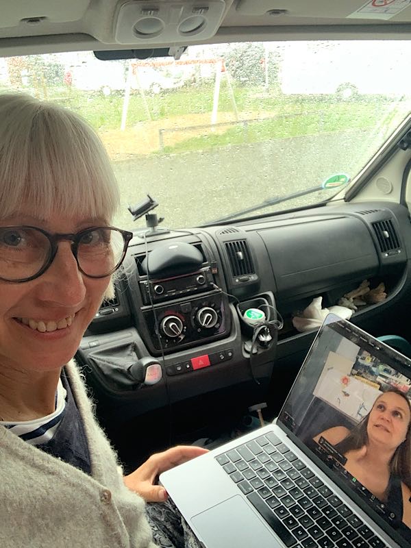 Frau im Wohnmobil bei einem Zoom-Call, Regen prasselt gegen die Windschutzscheibe, Gesprächspartnerin auf dem Laptop sichtbar.