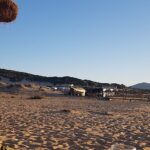 Frau mit hellem Haar sitzt unter einem Sonnenschirm am Strand und blickt in die Ferne, dahinter Dünenlandschaft und Abendlicht.