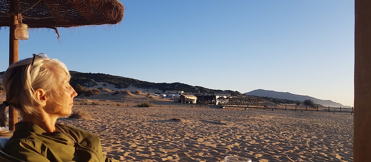 Frau mit hellem Haar sitzt unter einem Sonnenschirm am Strand und blickt in die Ferne, dahinter Dünenlandschaft und Abendlicht.