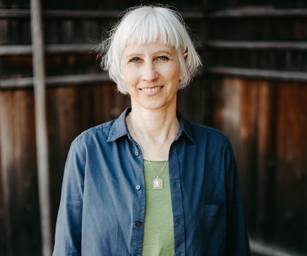 Frau mit weißem Pagenkopf blauer Bluse und grünem Shirt darunter vor dunkler Holzwand