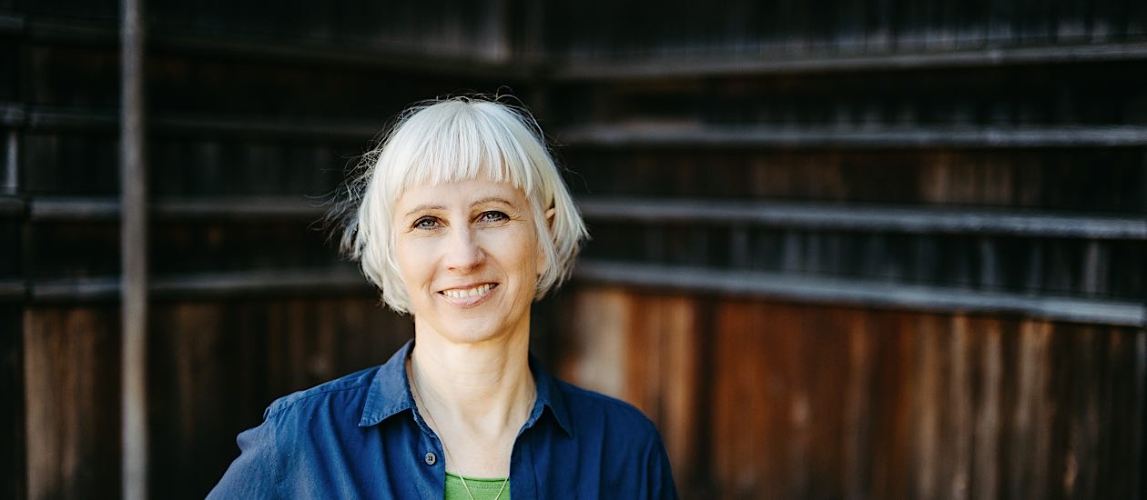 Frau mit weißem Pagenkopf blauer Bluse und grünem Shirt darunter vor dunkler Holzwand