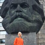 Weiße Frau im orangefarbenen Pullover steht vor dem Karl-Marx-Monument in Chemnitz, Blick nach oben, monumentaler Kopf im Hintergrund.