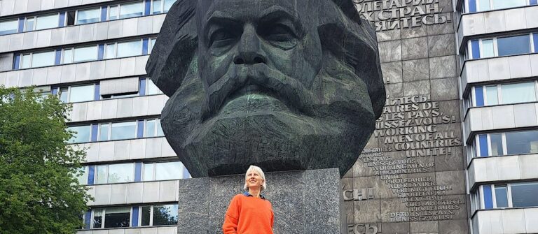 Weiße Frau im orangefarbenen Pullover steht vor dem Karl-Marx-Monument in Chemnitz, Blick nach oben, monumentaler Kopf im Hintergrund.