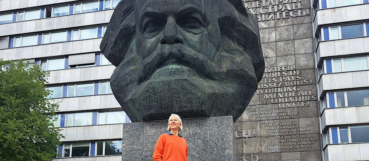Weiße Frau im orangefarbenen Pullover steht vor dem Karl-Marx-Monument in Chemnitz, Blick nach oben, monumentaler Kopf im Hintergrund.