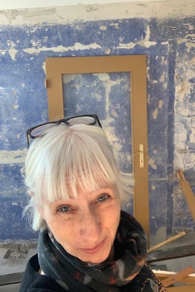 „Porträt einer Frau mit weißem Haar und Brille auf dem Kopf, aufgenommen in einem Raum mit freigelegter blauer Wand und Baustellenmaterial.“