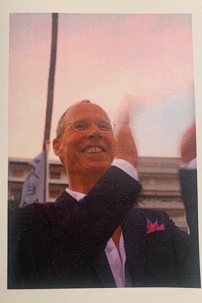 Fotografie eines Mannes im dunklen Anzug, der lächelnd applaudiert, aufgenommen im Freien vor einem Gebäude und Fahnen.