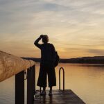 Frau steht am Ende eines Holzstegs am See und blickt in den Sonnenuntergang – Sinnbild für eine anstehende Entscheidung.