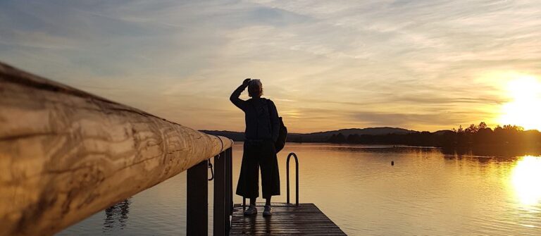 Frau steht am Ende eines Holzstegs am See und blickt in den Sonnenuntergang – Sinnbild für eine anstehende Entscheidung.