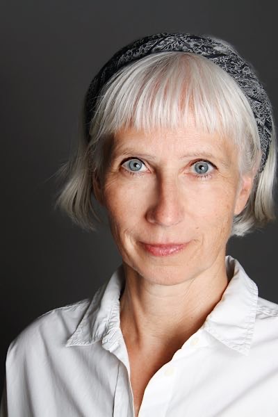 Porträt einer Frau mit grauem Haar, Pony und gemustertem Haarband, in weißer Bluse vor grauem Hintergrund, freundlich in die Kamera blickend.