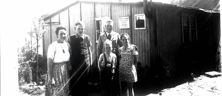 Schwarz-weißes Bild aus den späten 1940er Jahren zeigt eine Familie mit drei Kindern vor einer Baracke