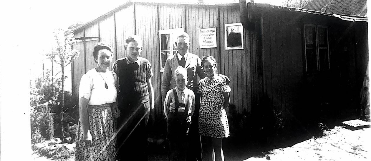 Schwarz-weißes Bild aus den späten 1940er Jahren zeigt eine Familie mit drei Kindern vor einer Baracke