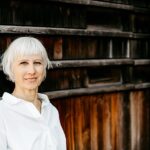 Birgit Krüger steht mit weißer Bluse und weißem Haar lächelnd an einer dunklen Holzfassade und blickt ruhig in die Kamera.