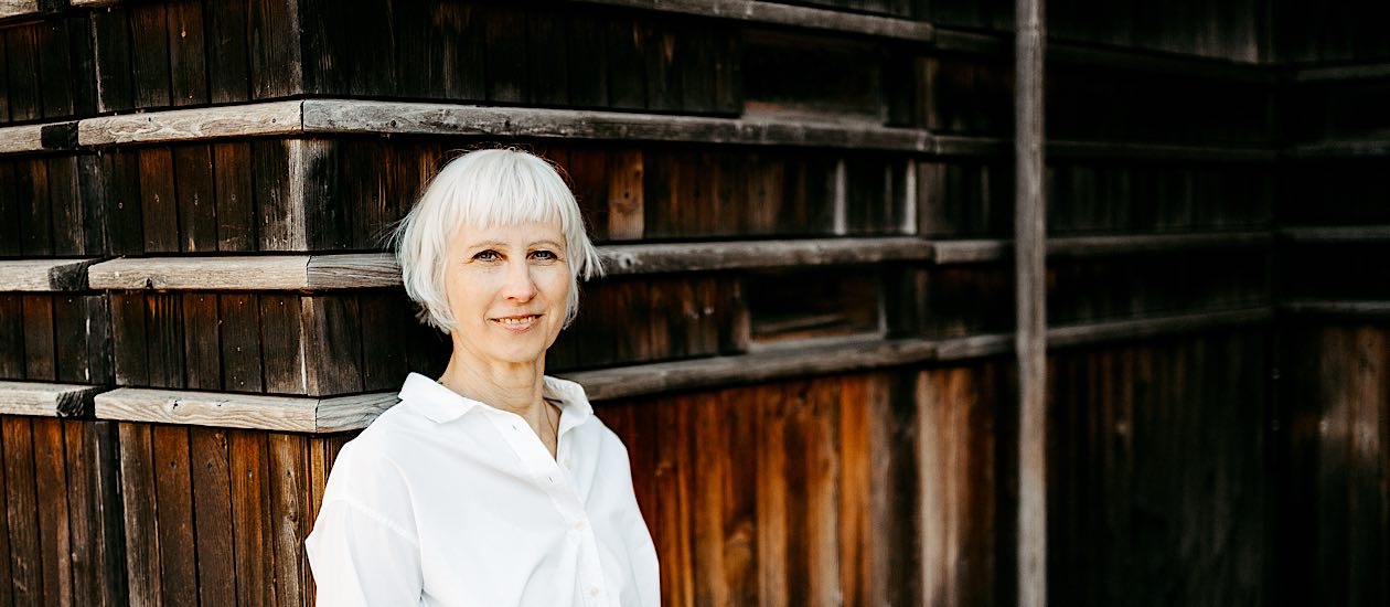 Birgit Krüger steht mit weißer Bluse und weißem Haar lächelnd an einer dunklen Holzfassade und blickt ruhig in die Kamera.