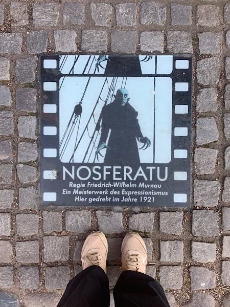 Gedenkplatte in Wismar für den expressionistischen Stummfilm ‚Nosferatu‘ von 1921 – Blick auf das Filmrelief im Straßenpflaster mit den beigen Sneakern von Focusing-Coach Birgit Krüger im Bild