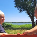 Zwei Frauen sitzen auf einer roten Bank im Grünen mit Blick auf einen See, Birgit Krüger (links) mit weißem Haar, im Gespräch mit Philine, einer Frau mit lockigem blondem Haar. Im Hintergrund Bäume, Wiese und Wasser unter blauem Himmel