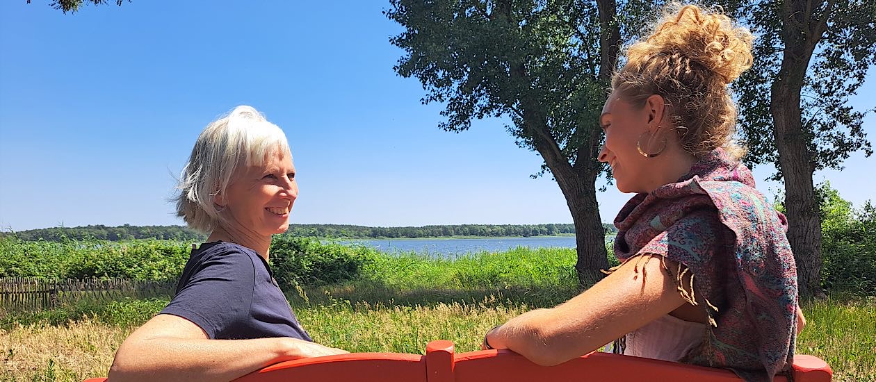 Zwei Frauen sitzen auf einer roten Bank im Grünen mit Blick auf einen See, Birgit Krüger (links) mit weißem Haar, im Gespräch mit Philine, einer Frau mit lockigem blondem Haar. Im Hintergrund Bäume, Wiese und Wasser unter blauem Himmel