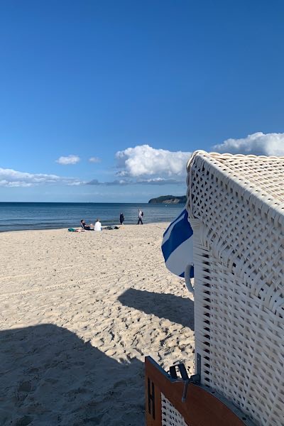 Strandkorb von der Seite mit blau weißem Sonnenschutz bei Sonnenschein auf weißem Strand mit Meer und Menschen im Vordergrund